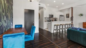 Ein modernes Wohnzimmer mit einem Holzesstisch, blauen Stühlen, einem türkisgrünen Sofa und einer Küchenzeile mit weißen Schränken und einem Barhocker.