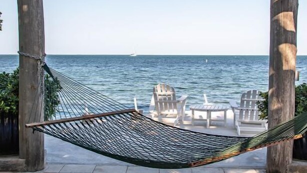 Eine Hängematte unter einer Holzpergola mit Blick auf einen Strand, mit Stühlen und einem Segelboot in der Ferne.