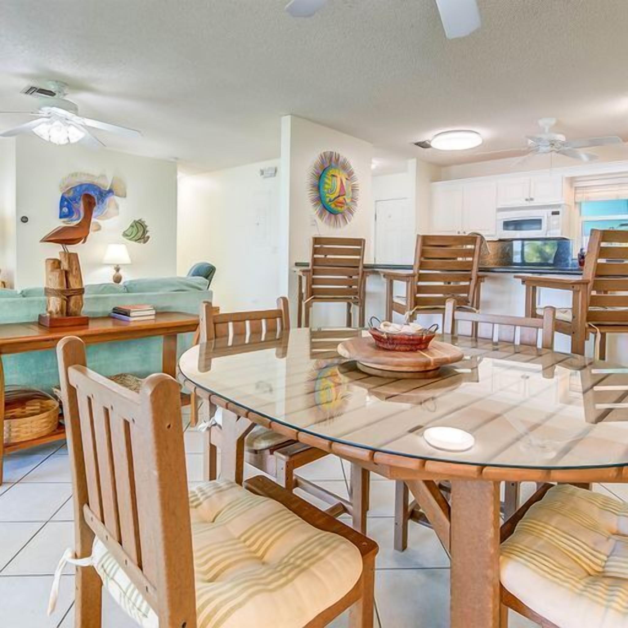 A dining area with a glass table surrounded by wooden chairs, a kitchen in the background, and a ceiling fan.