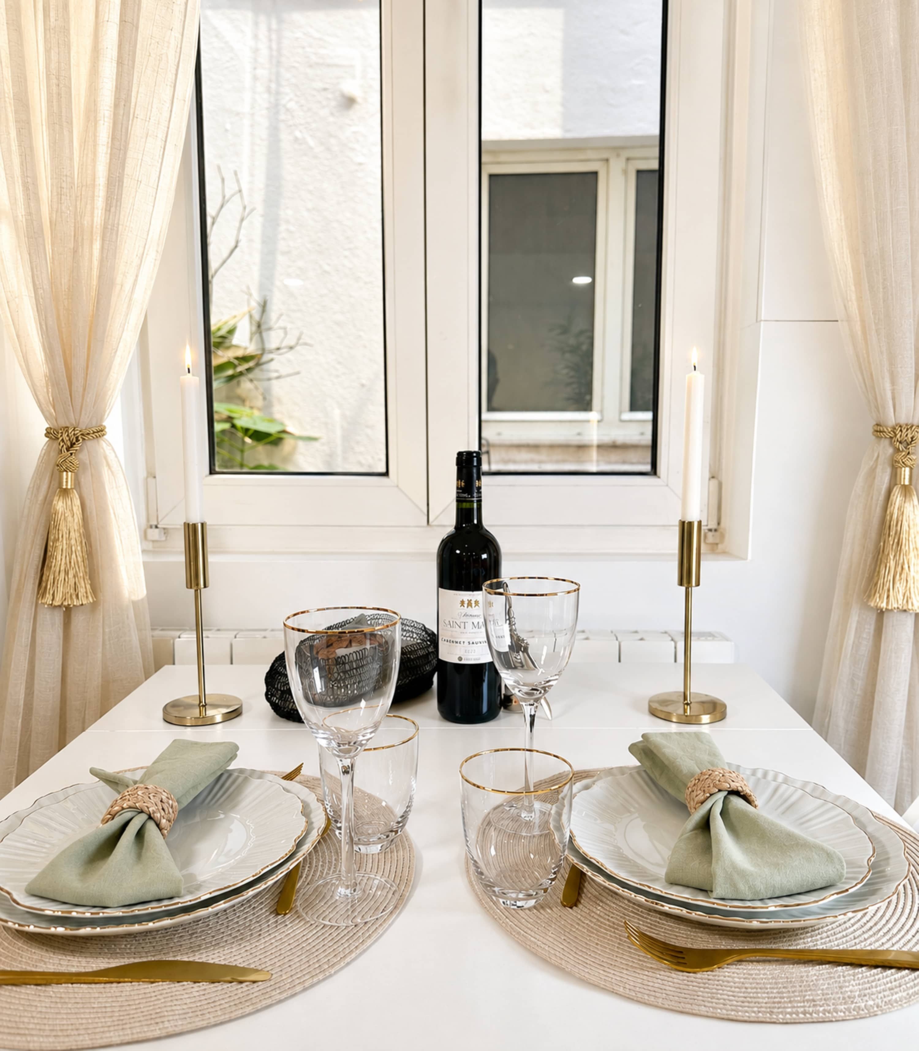 A dining table set with plates, glasses, a bottle, and candles, with a window and curtains in the background.
