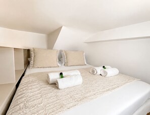 A neatly made bed with rolled towels and a bottle, a white wall, and a sloped ceiling. - Stylish studio near the Tuileries Gardens (Paris)