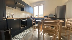 A modern kitchen with dark cabinets, a built-in oven, a dining table with chairs, and a large window providing natural light.