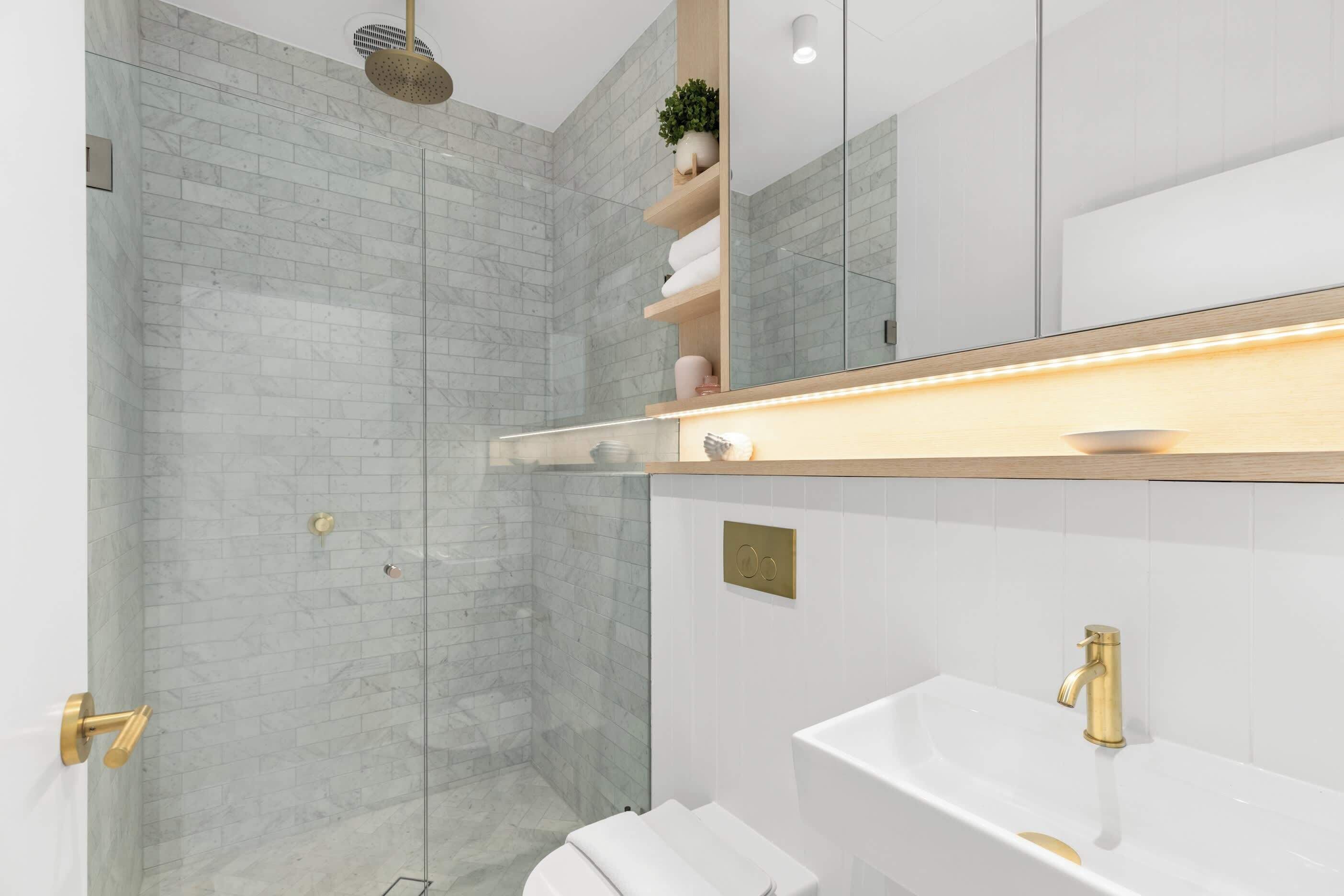 A modern bathroom with a glass shower enclosure, a white sink with a gold faucet, and a wooden shelf with towels.