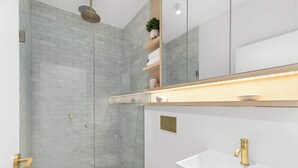 Un baño moderno con cabina de ducha de vidrio, lavabo blanco con grifo dorado y estante de madera con toallas.