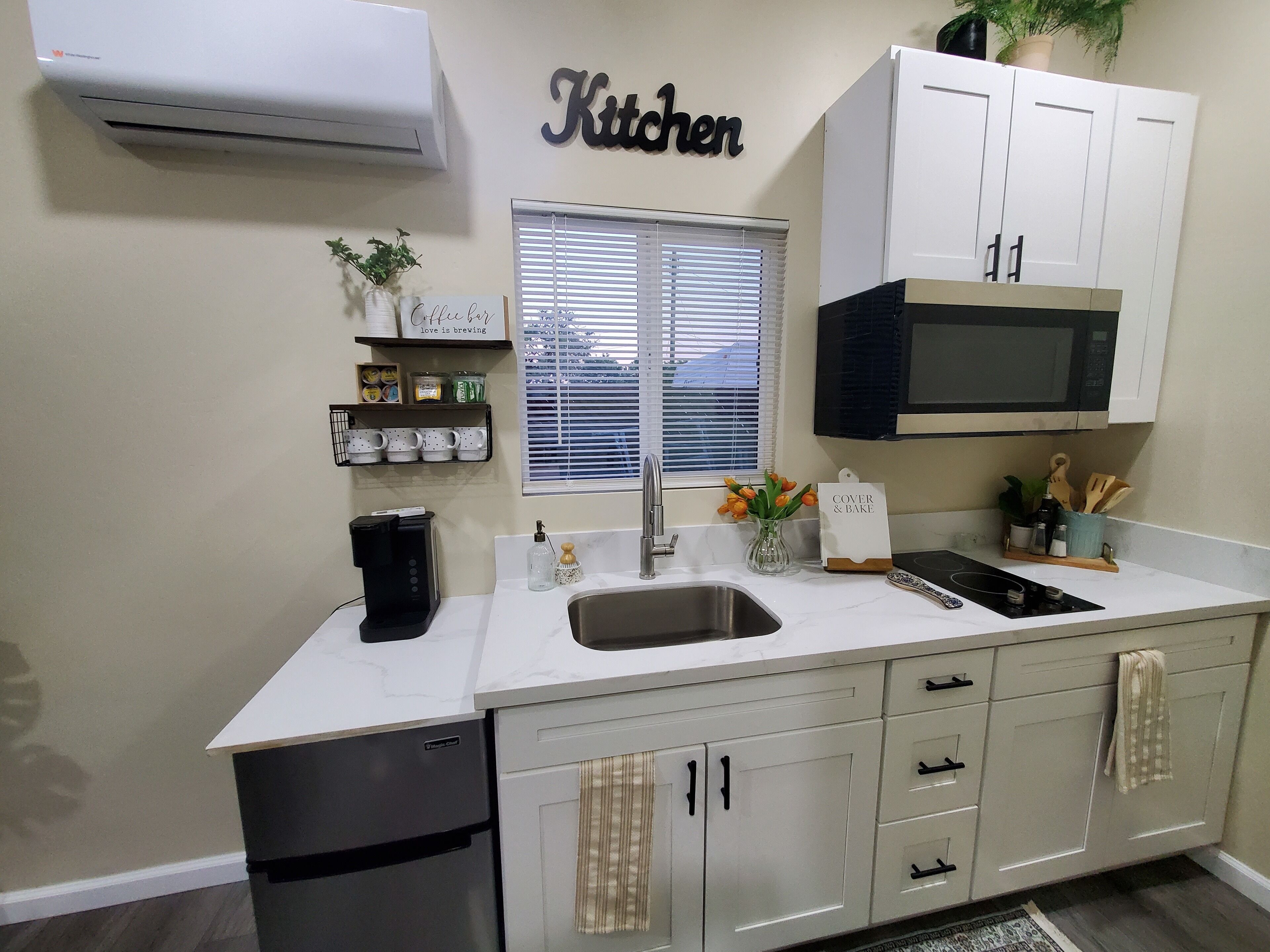 A modern kitchen with white cabinets, a black microwave, a coffee maker, and a window with blinds.