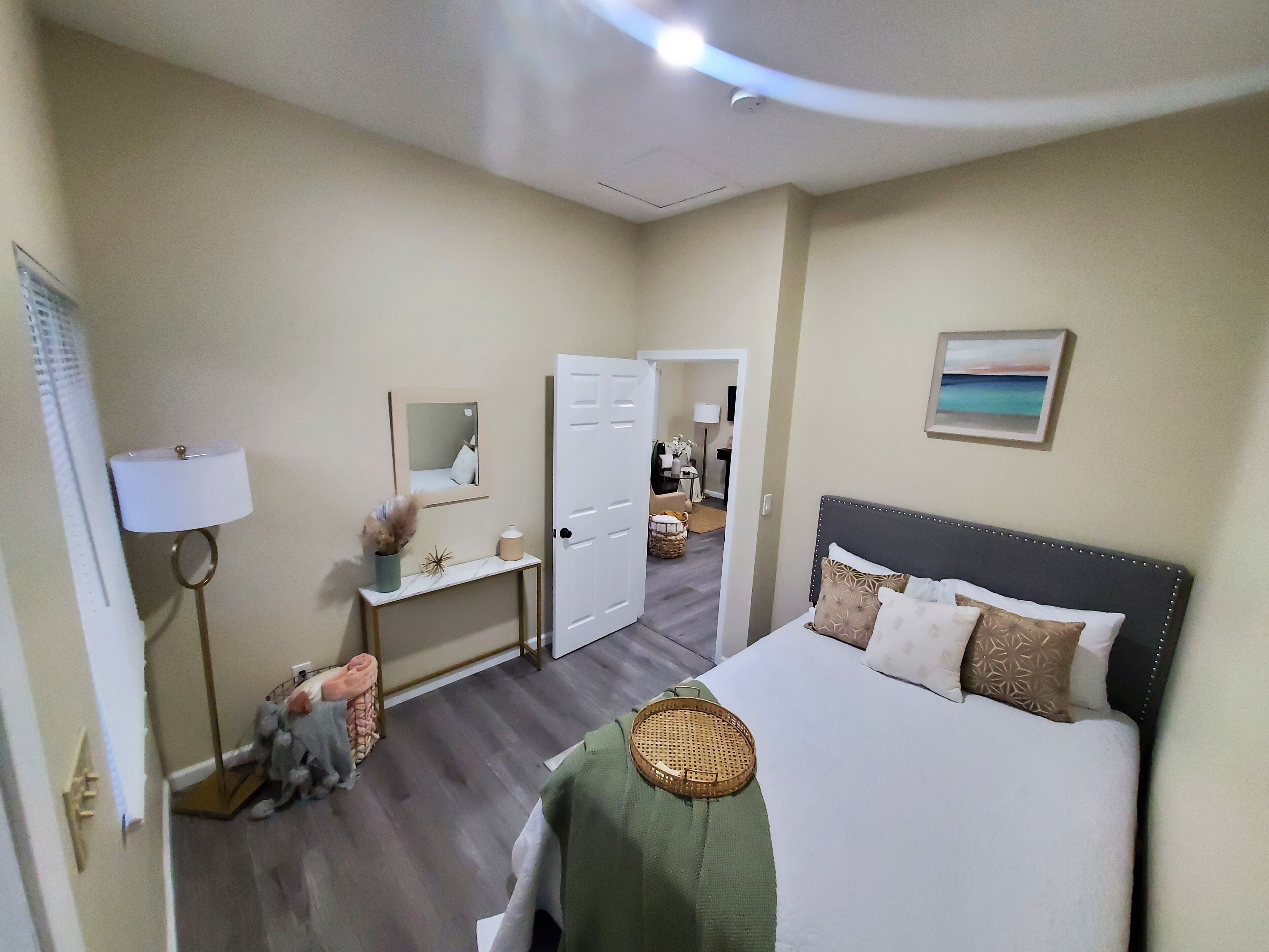 A hotel room with a bed, bedside table, lamp, mirror, and a painting on the wall.