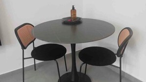 Une table ronde avec un pied noir et trois chaises noires à assise tressée. Une bouteille de gel hydroalcoolique est posée sur la table.