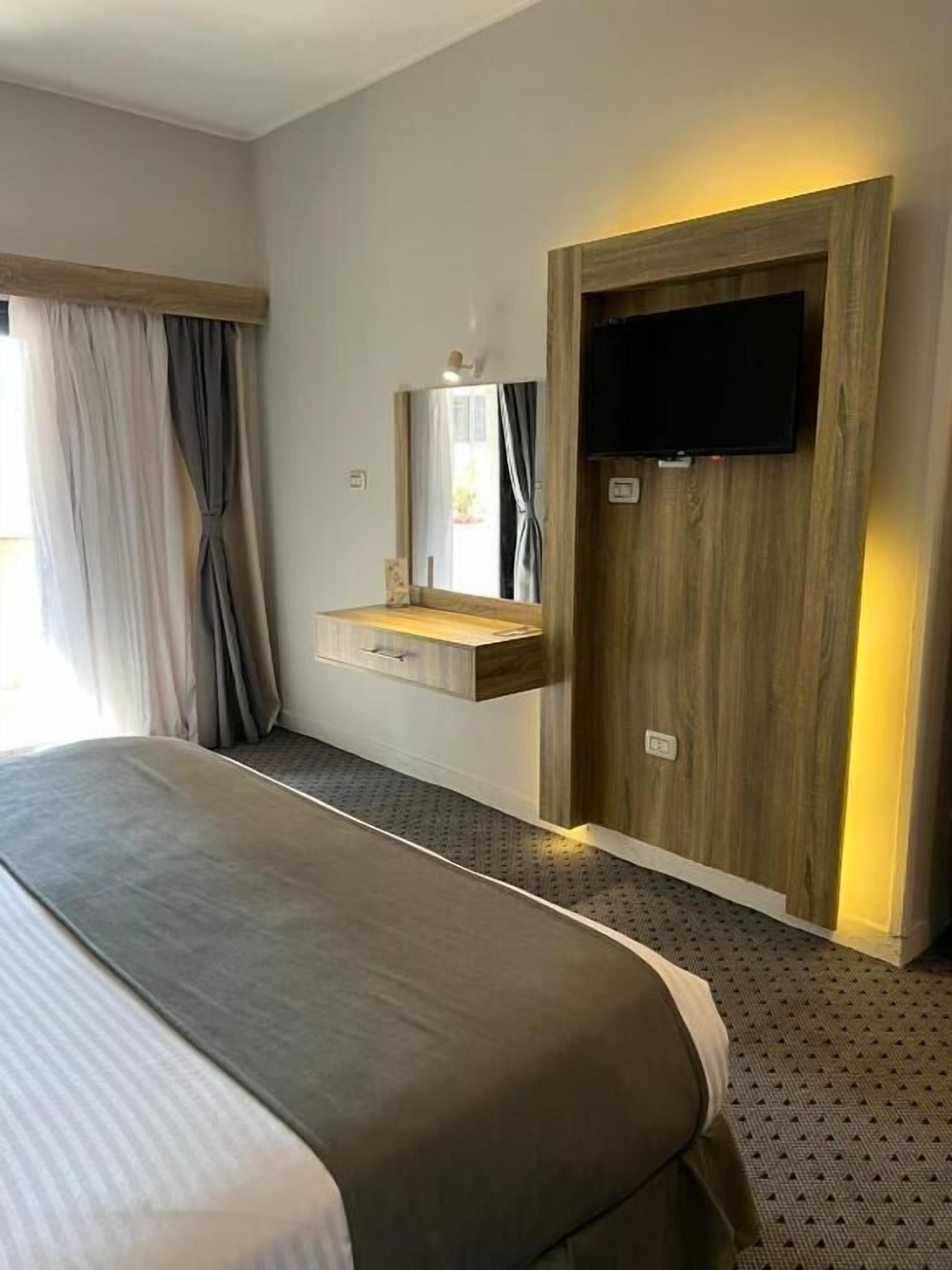 A hotel room with a bed, a wooden wardrobe, a mounted television, and a window with curtains.
