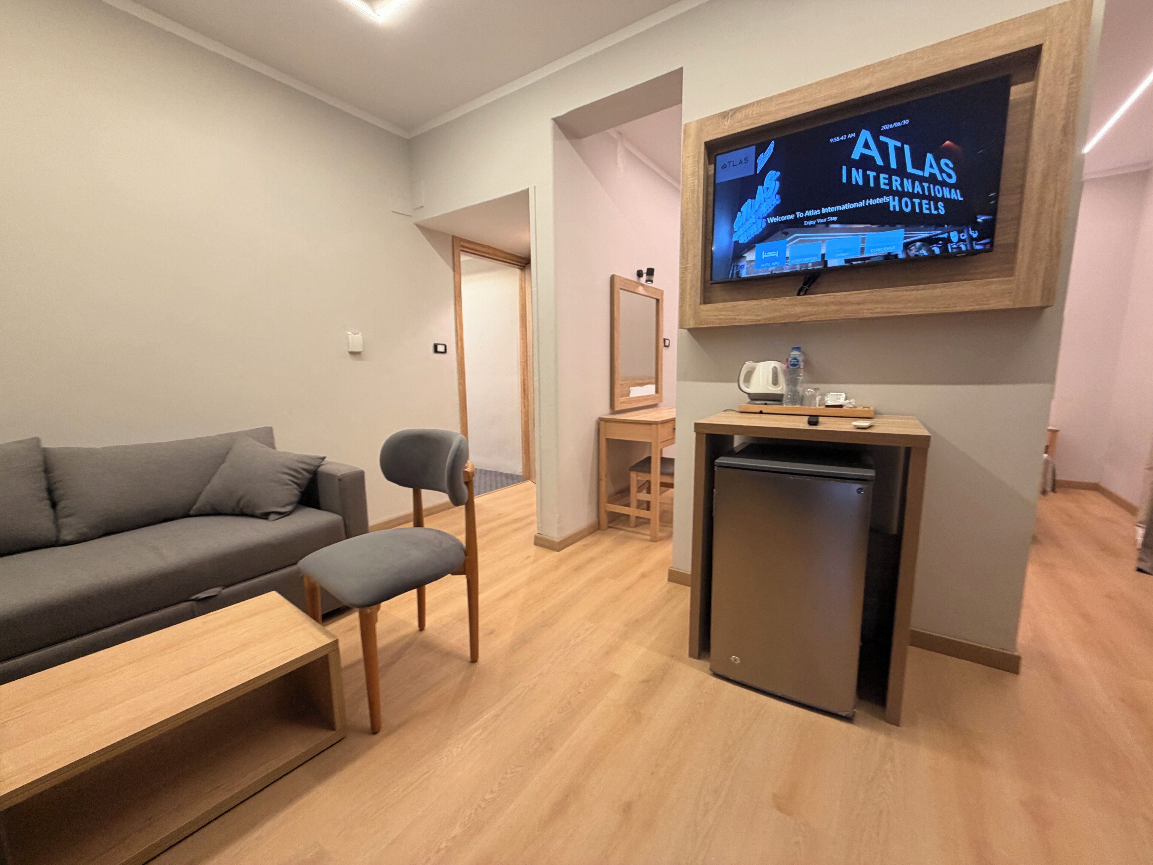 Un salon moderne avec un canapé gris, une table basse en bois, un petit téléviseur fixé au mur, un réfrigérateur compact et une chaise en bois.