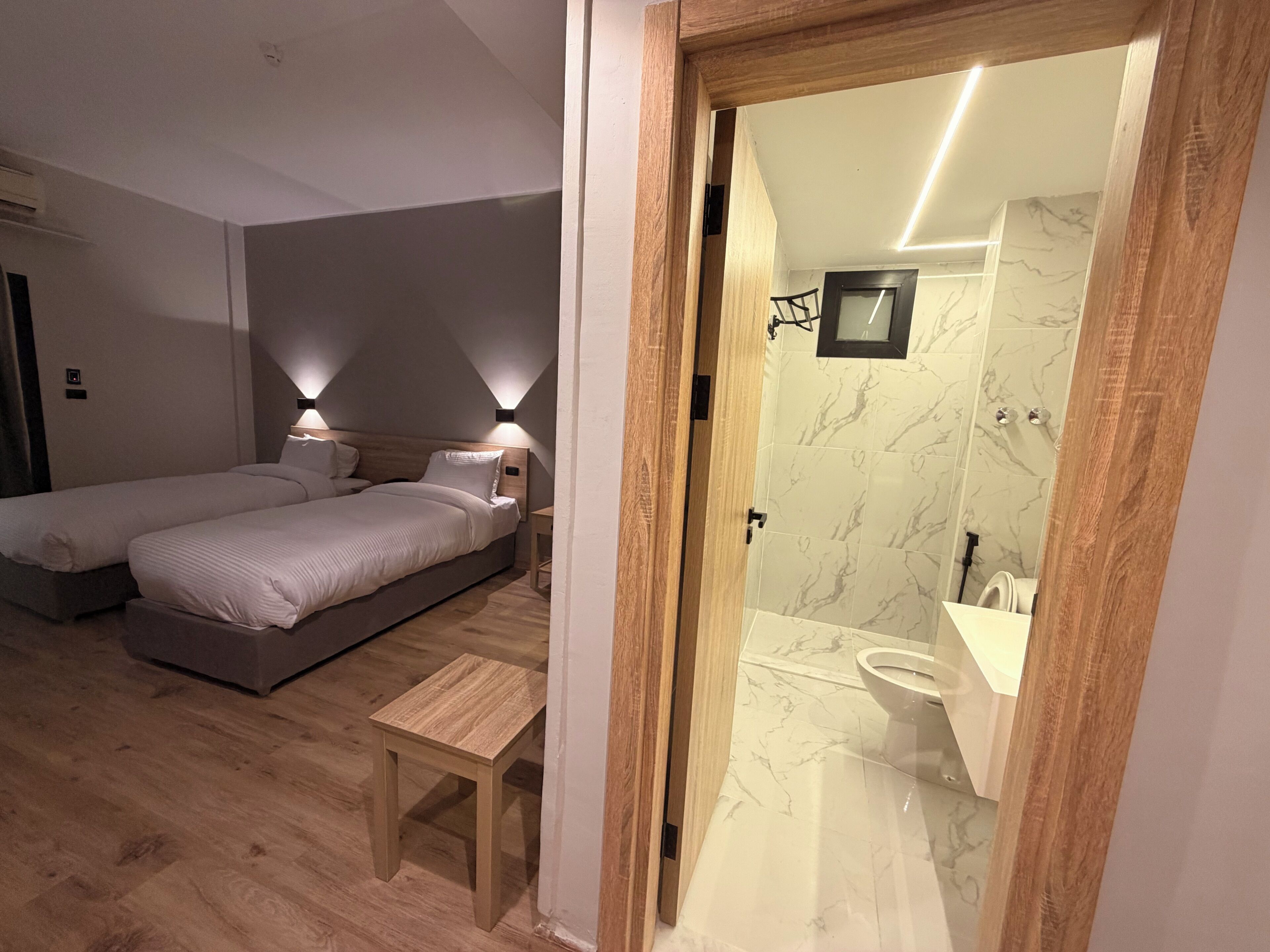 A bedroom with two beds, a wooden bench, and a bathroom with a marble floor and a mirror.