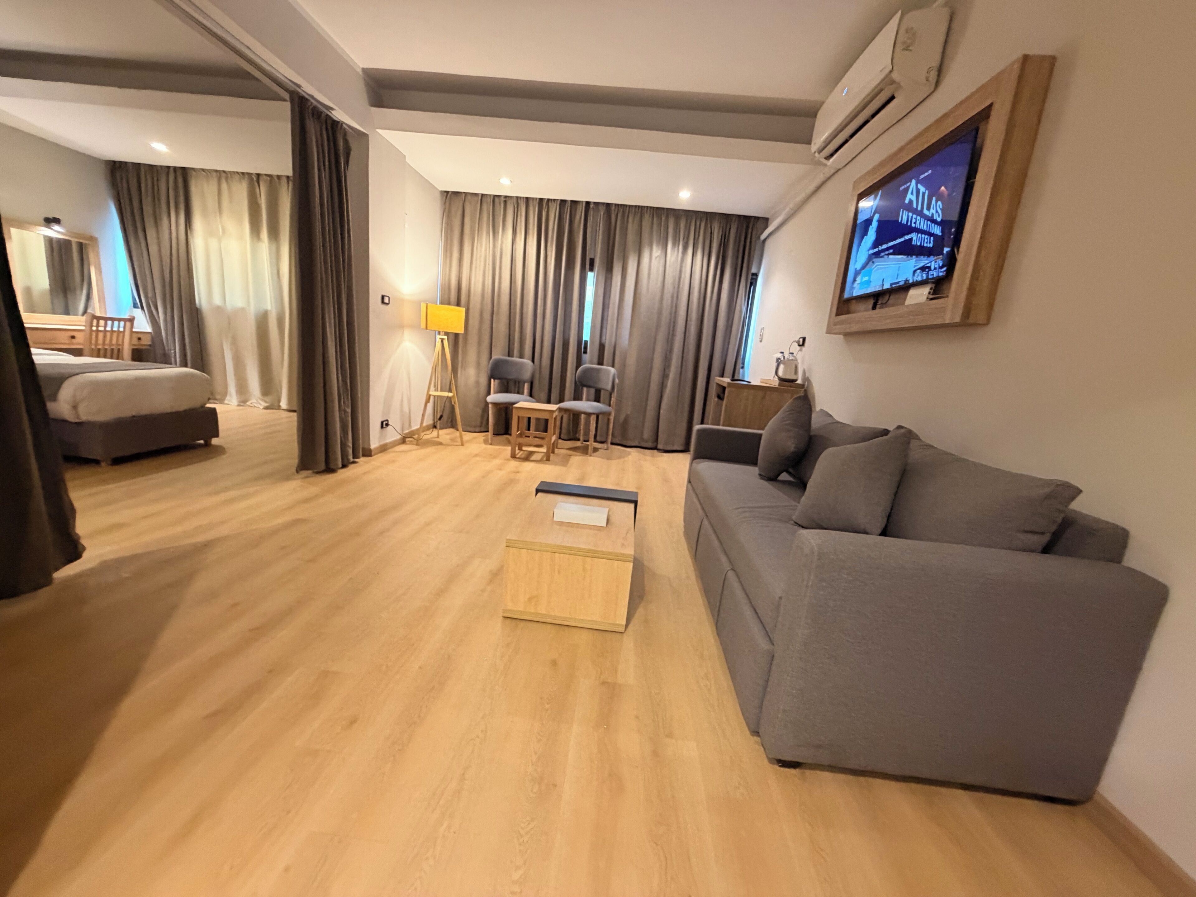 A modern hotel room with a grey sofa, a wooden coffee table, a desk with a chair, a bed, and a television mounted on the wall.