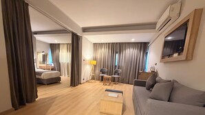 A modern hotel room with a wooden floor, a grey sofa, a small wooden table, a TV mounted on the wall, and a bed with curtains. - Atlas Hotels (Cairo)