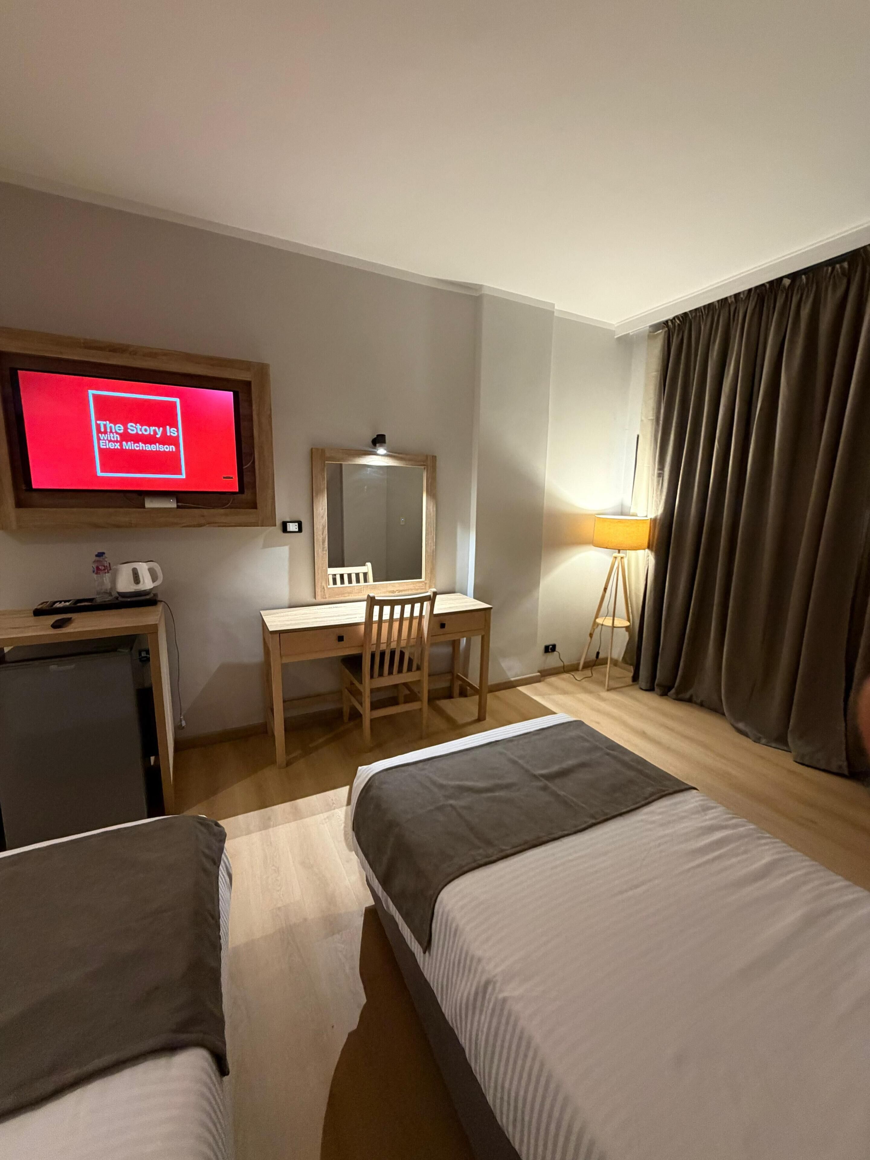 Ein Hotelzimmer mit zwei Betten, einem Schreibtisch, einem Fernseher, einem Spiegel, einer Lampe und einem fenster mit Vorhängen.