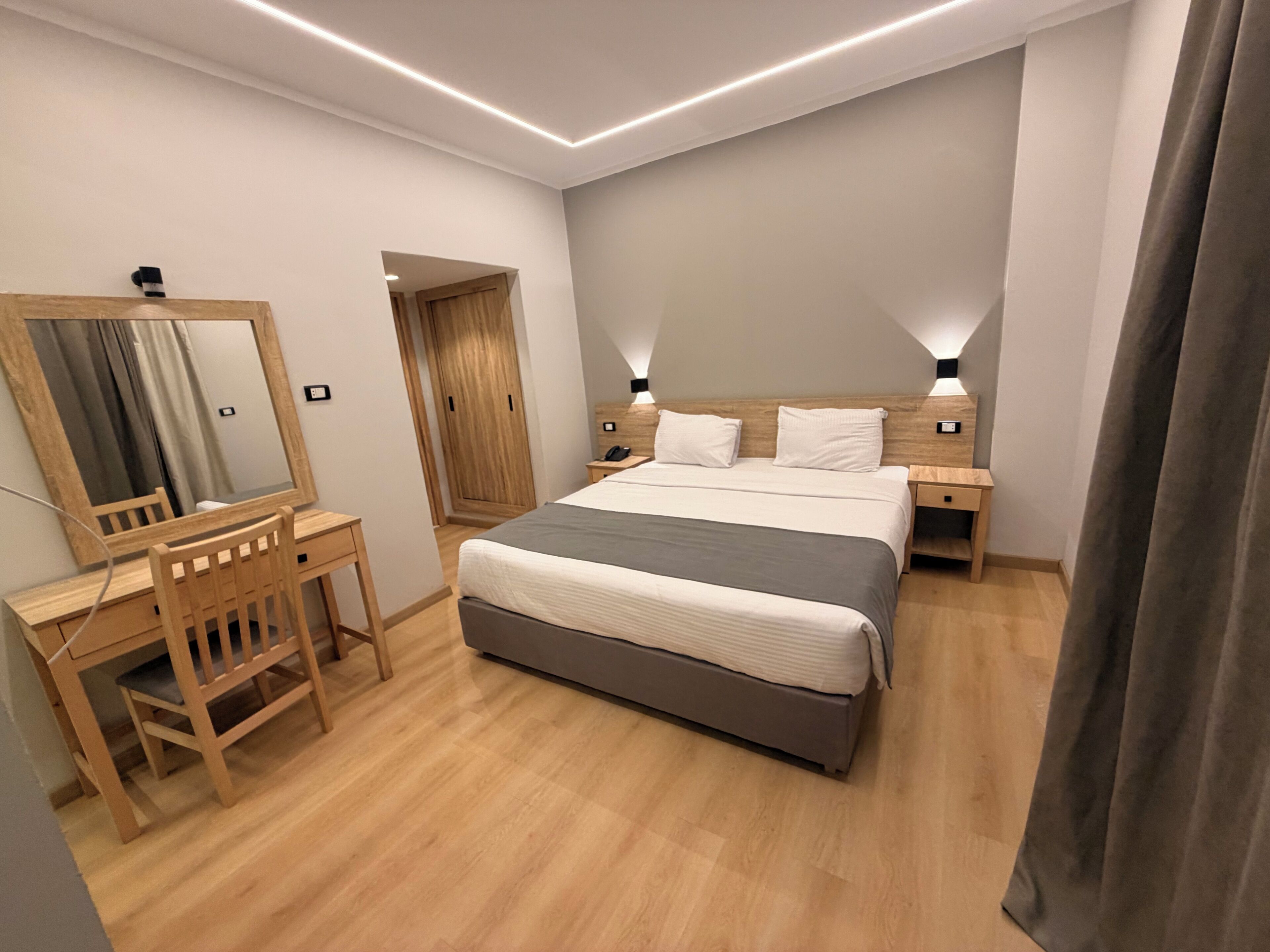 A modern bedroom with a wooden desk, a bed with a grey headboard, bedside tables with lamps, and a wooden wardrobe.