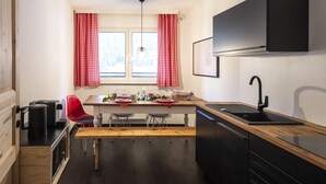 Een kleine keuken met een houten tafel, rood-wit geruit gordijn, een zwarte spoelbak en een zwart kastje boven het aanrecht.