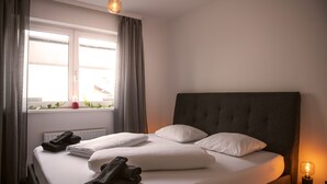 Een netjes opgemaakt bed met witte lakens en donkere handdoeken, een raam met gordijnen, en een bedlampje.