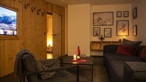 Een gezellige woonkamer met een houten wand, een aan de muur bevestigde televisie, een bank, een stoel, een bijzettafeltje met een kaars en glazen, en lijstjes met foto’s aan de wand.