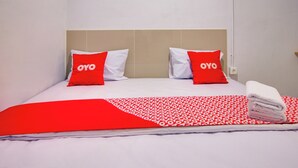 Ein Hotelzimmer mit einem Bett, zwei rote Kissen mit dem Schriftzug "OYO", einer rot-weißen Bettdecke und einem ordentlich gefalteten weißen Handtuch.