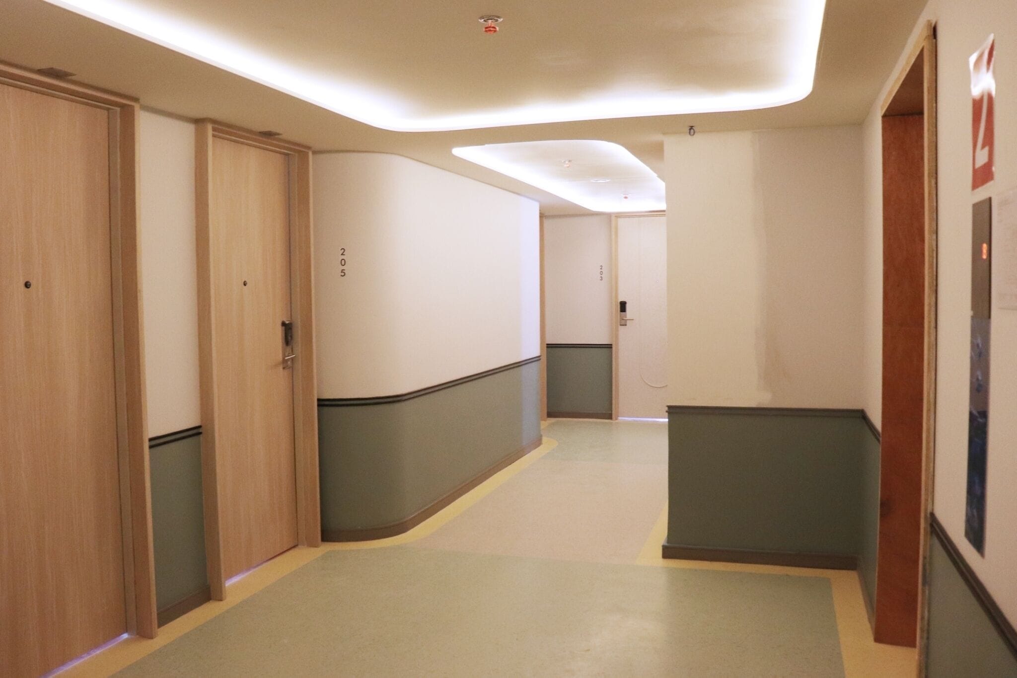 Een gang met houten deuren, groenbeige wanden en een lichtgekleurde vloer.
