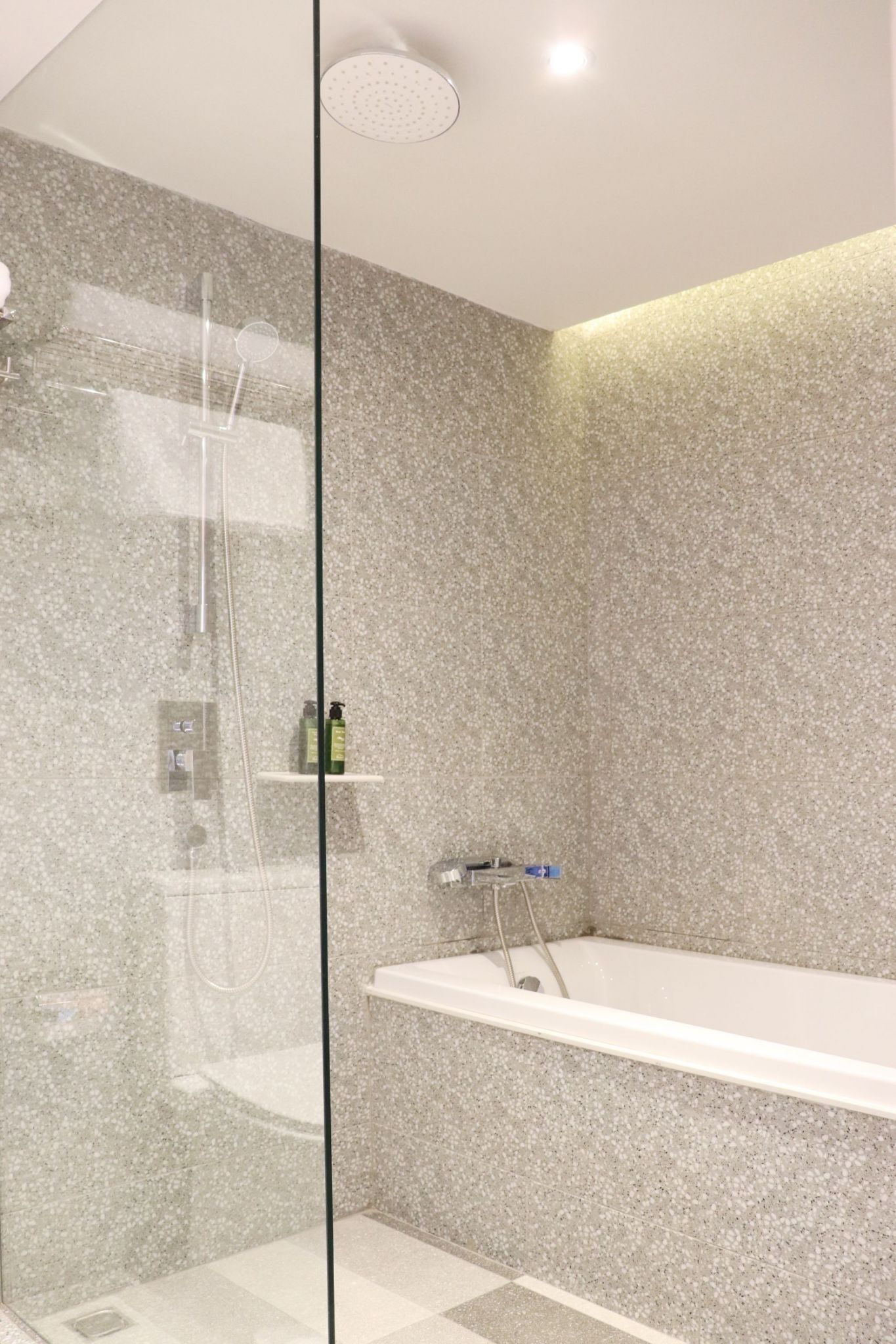 A modern bathroom with a glass-enclosed shower, a white bathtub, and a granite-tiled wall.