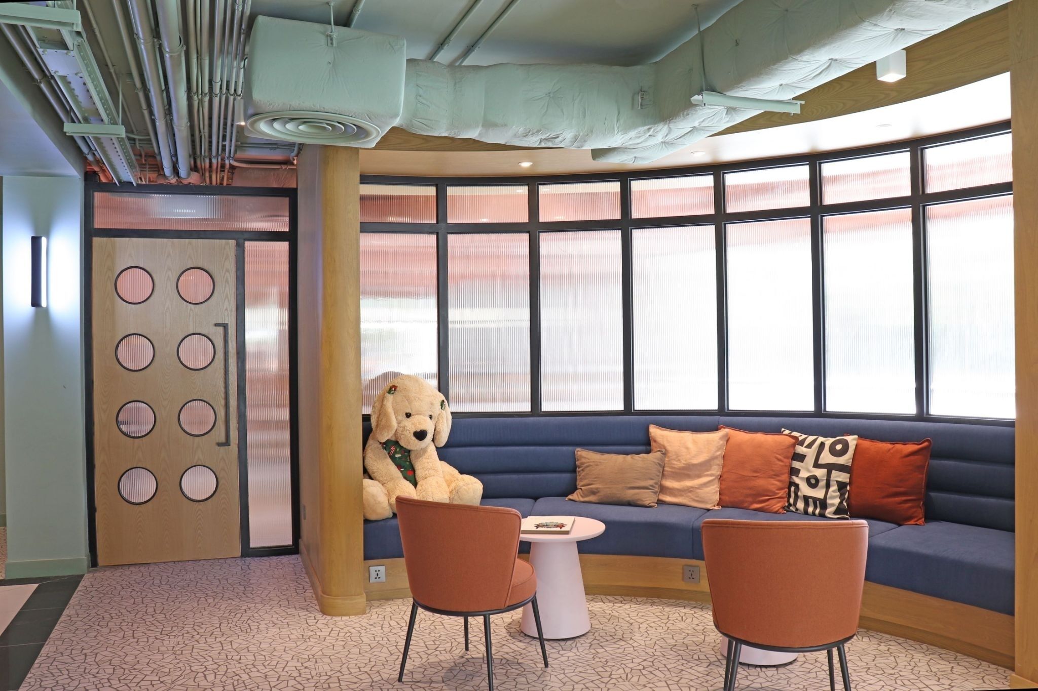 A modern lounge area with a blue sofa, orange chairs, and a large teddy bear.