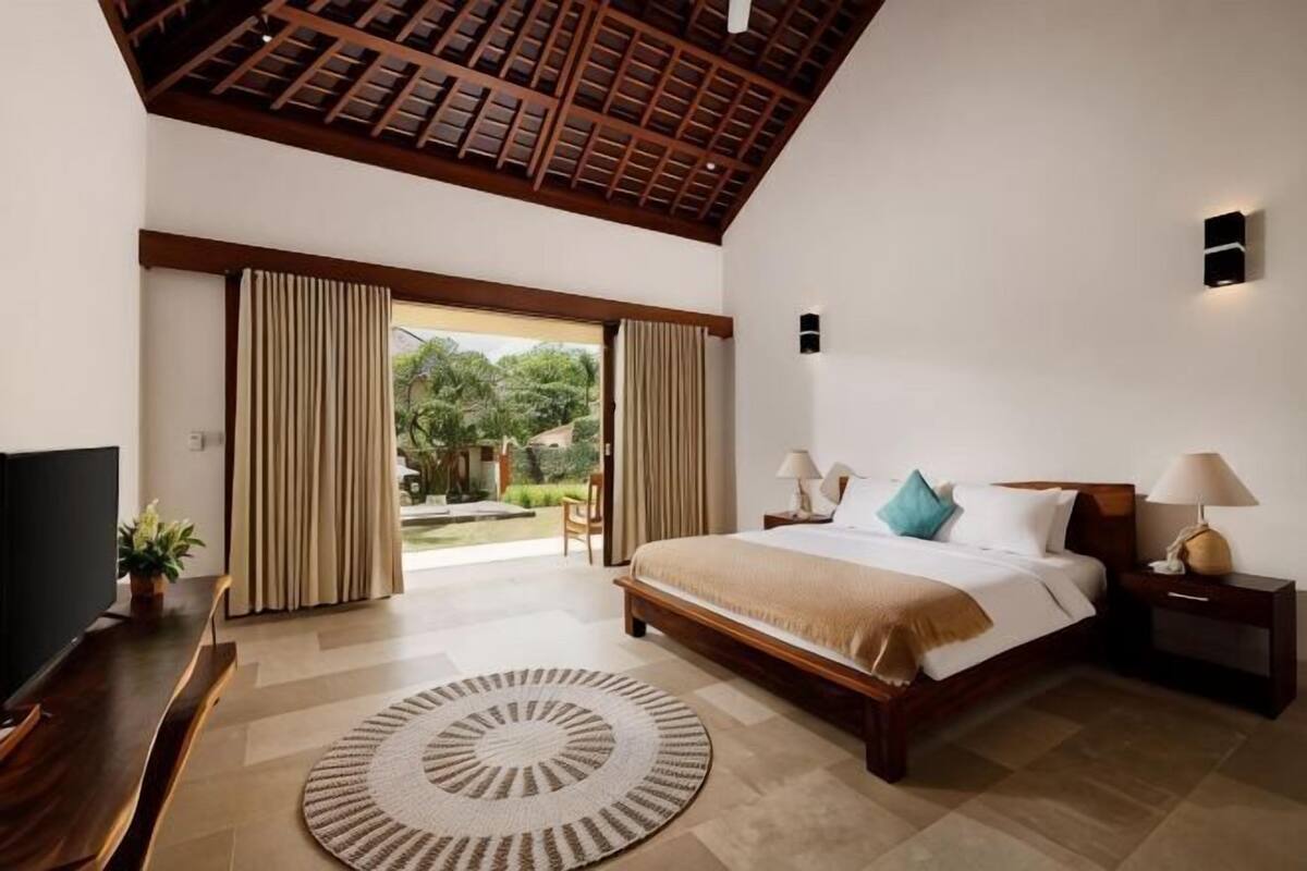 Un dormitorio con cama, mesitas de noche, televisor, alfombra circular y vista por una puerta.