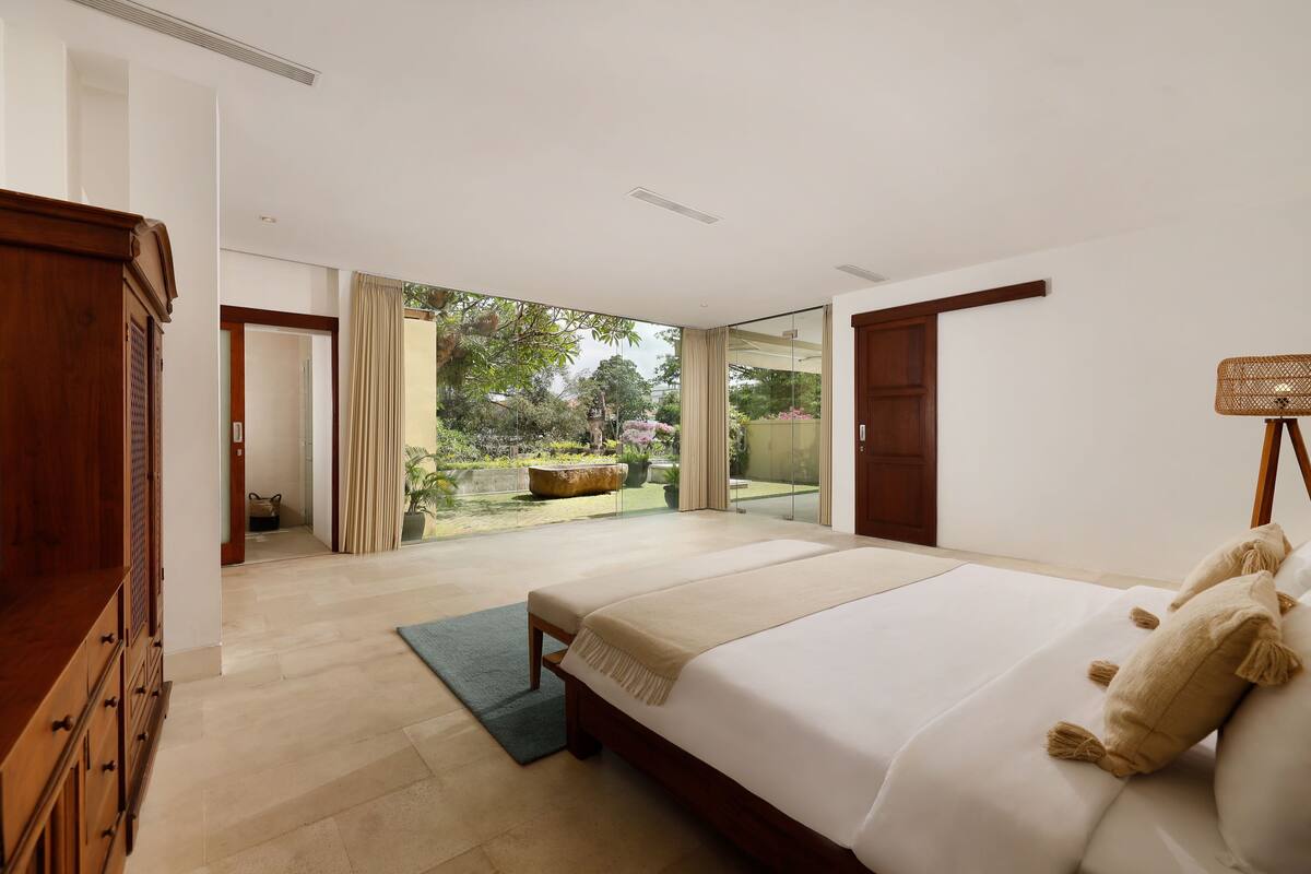 Un dormitorio amplio con una cama grande, un armario de madera y vistas a un jardín a través de un ventanal.