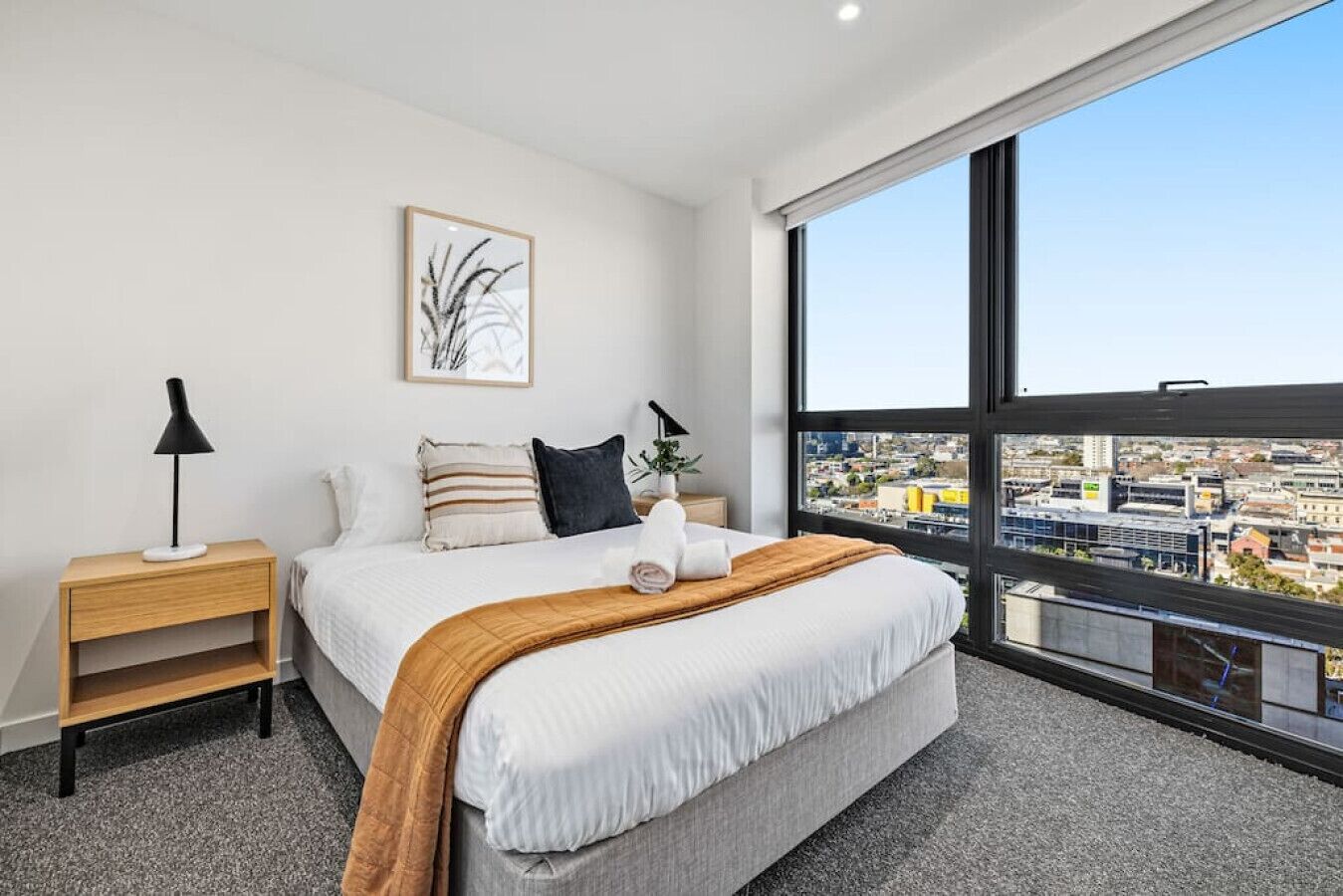 Une chambre d’hôtel moderne avec un grand lit, deux tables de chevet, une vue sur la ville grâce à de grandes fenêtres et une œuvre d’art encadrée au mur.