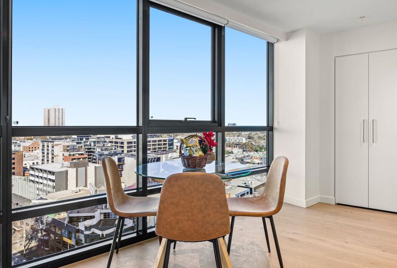 Una habitación moderna con ventanas amplias, una mesa de vidrio, dos sillas y vistas a la ciudad.