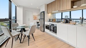 A modern kitchen with white cabinetry, a dining area with a glass table, and a view of the city through large windows. - Spectacular Views, Comfort, Convenience. Gym, Pool, Rooftop, Tram, walk to Crown (Southbank)