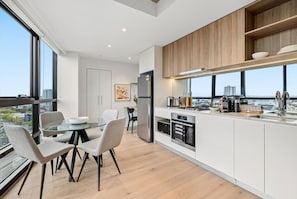 Une cuisine moderne avec des meubles blancs, un coin repas doté d’une table en verre et une vue sur la ville grâce à de grandes fenêtres.