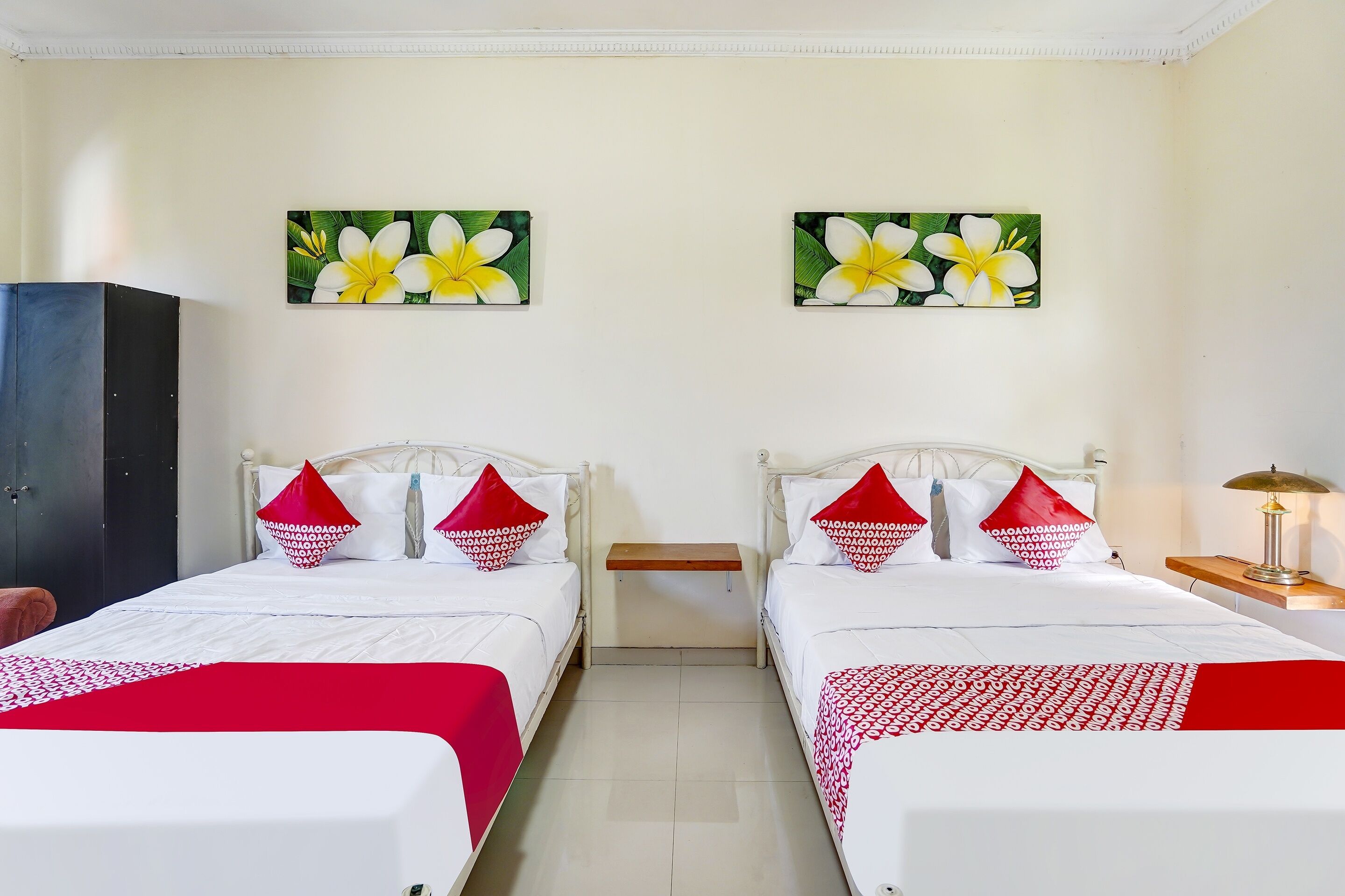 A hotel room with two beds, each with white bedding and red accents, a nightstand, a lamp, and two wall-mounted paintings of flowers.