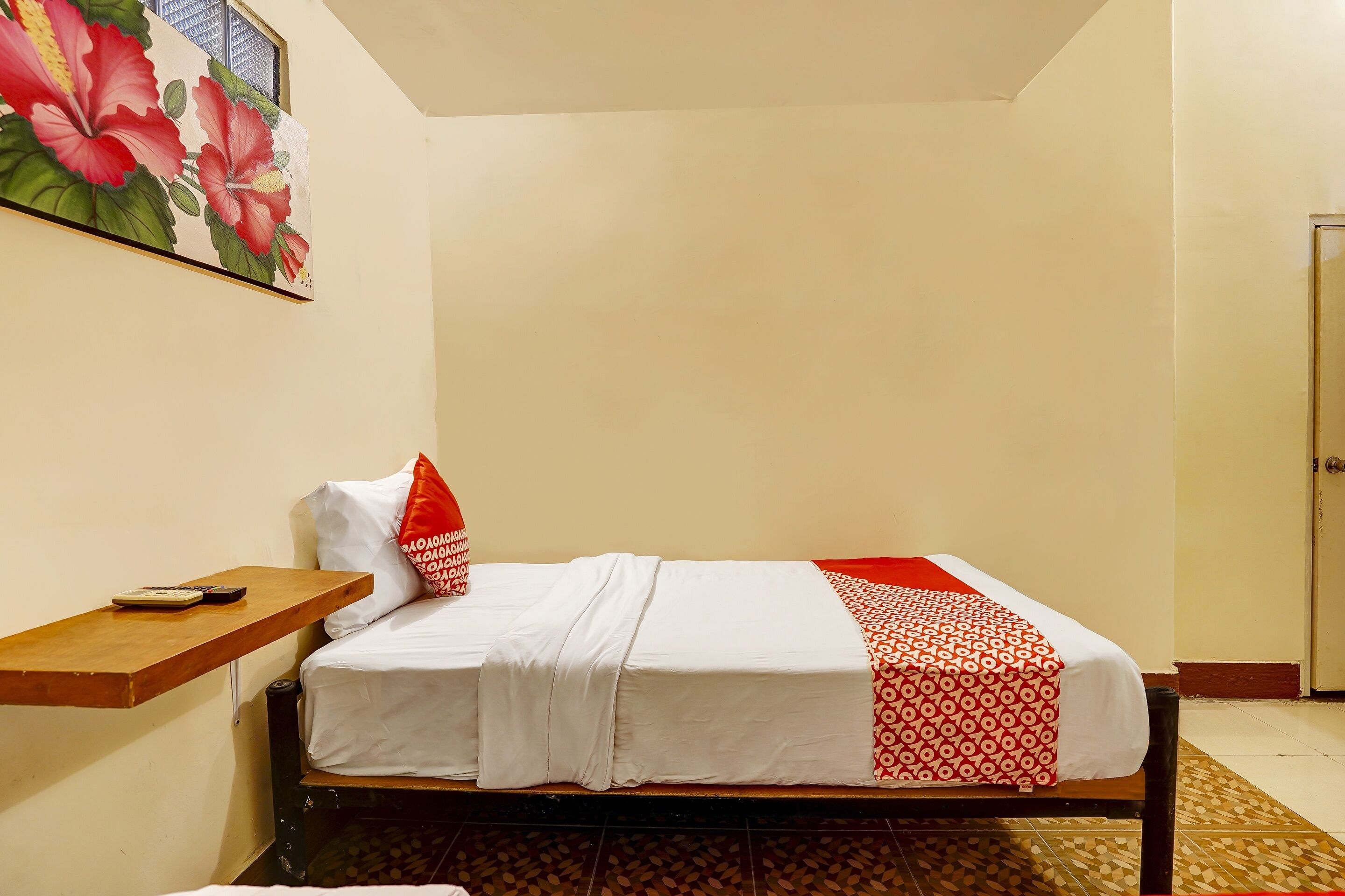 A single bed with white bedding and a red and white patterned throw, a small wooden shelf with a phone and keys, and a floral painting on the wall.