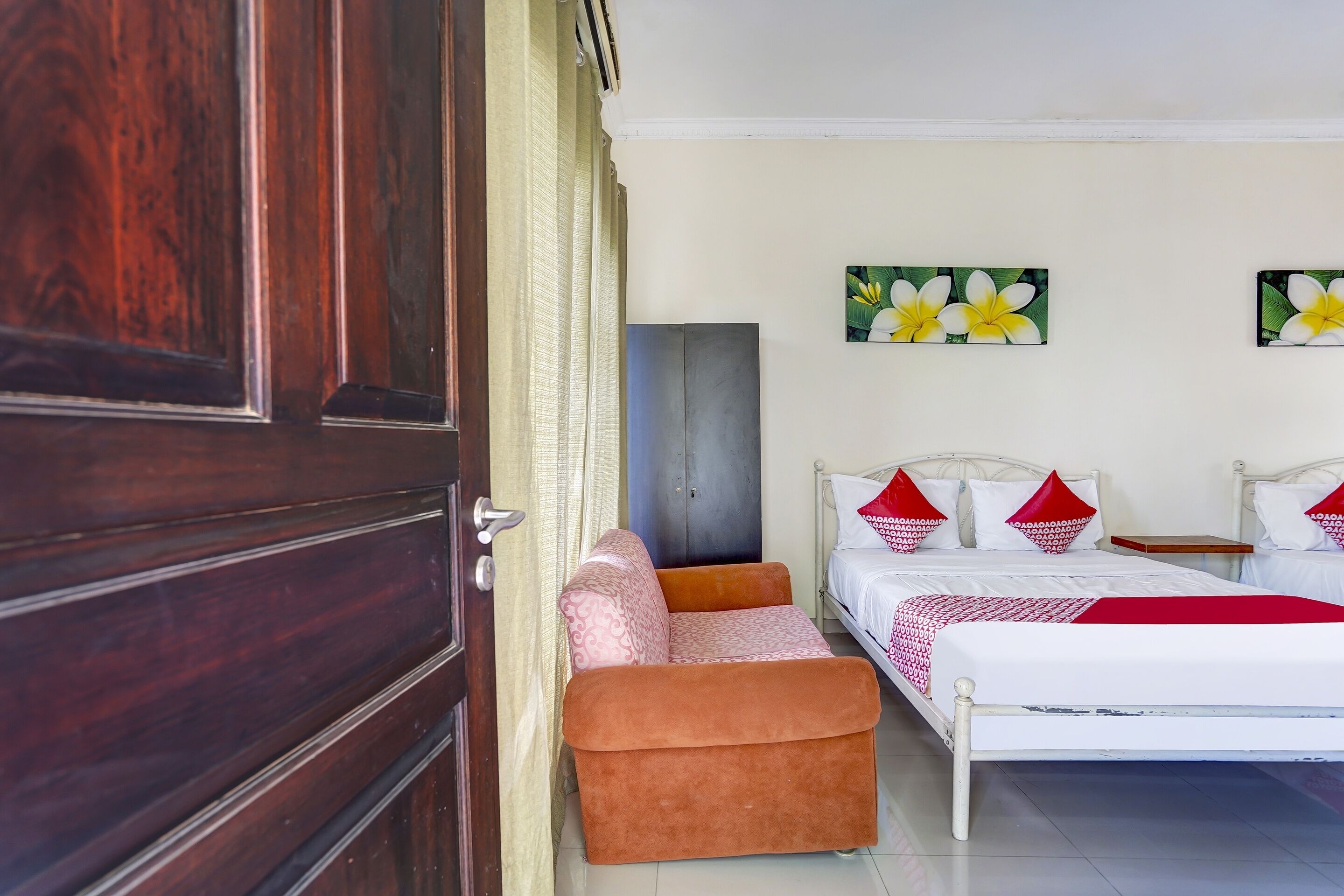 A bedroom with a wooden door, a bed with red and white bedding, a bench, and two wall-mounted flower paintings.