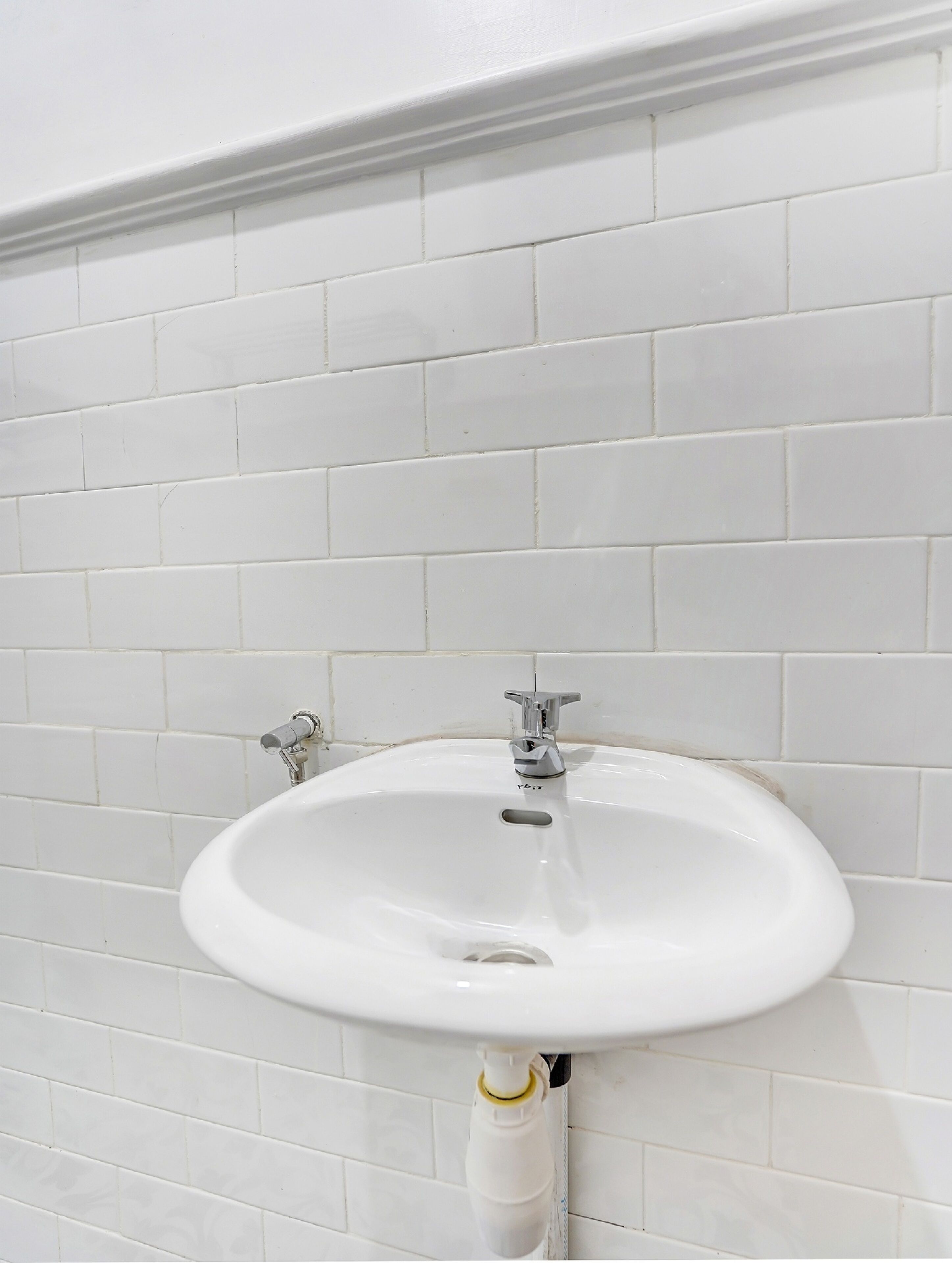 Ein Badezimmer mit einem weißen Waschbecken, zwei Wasserhähnen und einer weißen, gefliesten Wand.