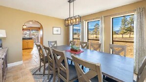 A dining area with a long table, chairs, and a chandelier. There is a view of trees and mountains through large windows. - Flagstaff Family Retreat~Game Room~Hot Tubs (Flagstaff)
