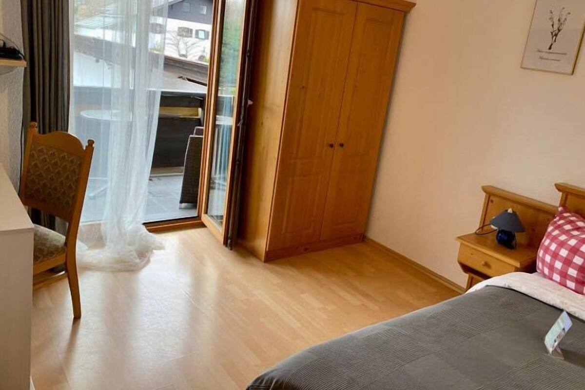 Un dormitorio con cama, armario de madera, silla, mesita de noche y ventana con cortinas.
