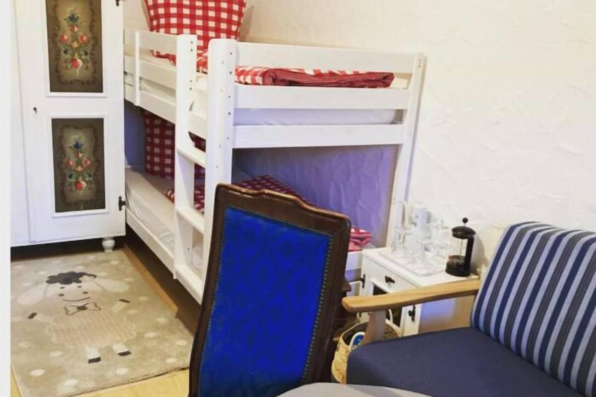 Una cama litera con una colcha a cuadros rojo y blanco, una silla azul con cojín y un control remoto sobre una mesa.