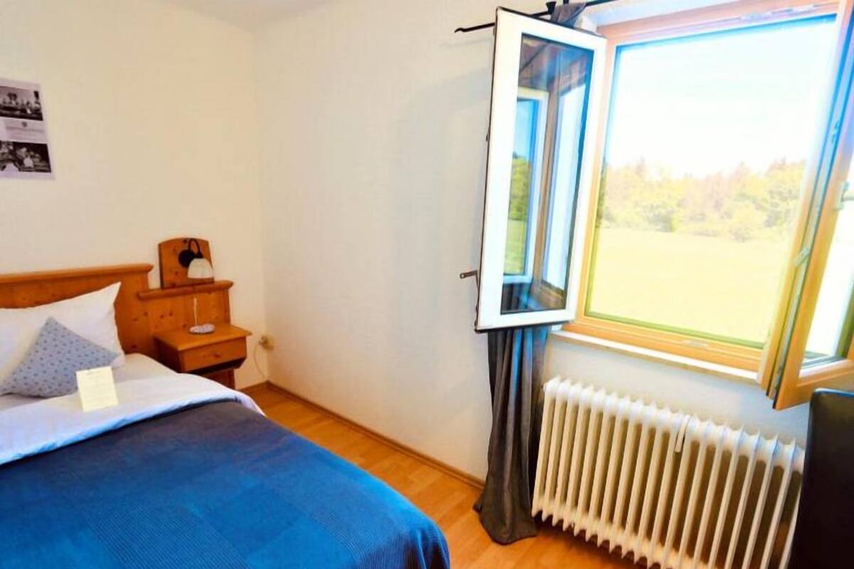 Un dormitorio con cama, mesita de noche, cómoda, ventana y vistas al verdor, además de un radiador.