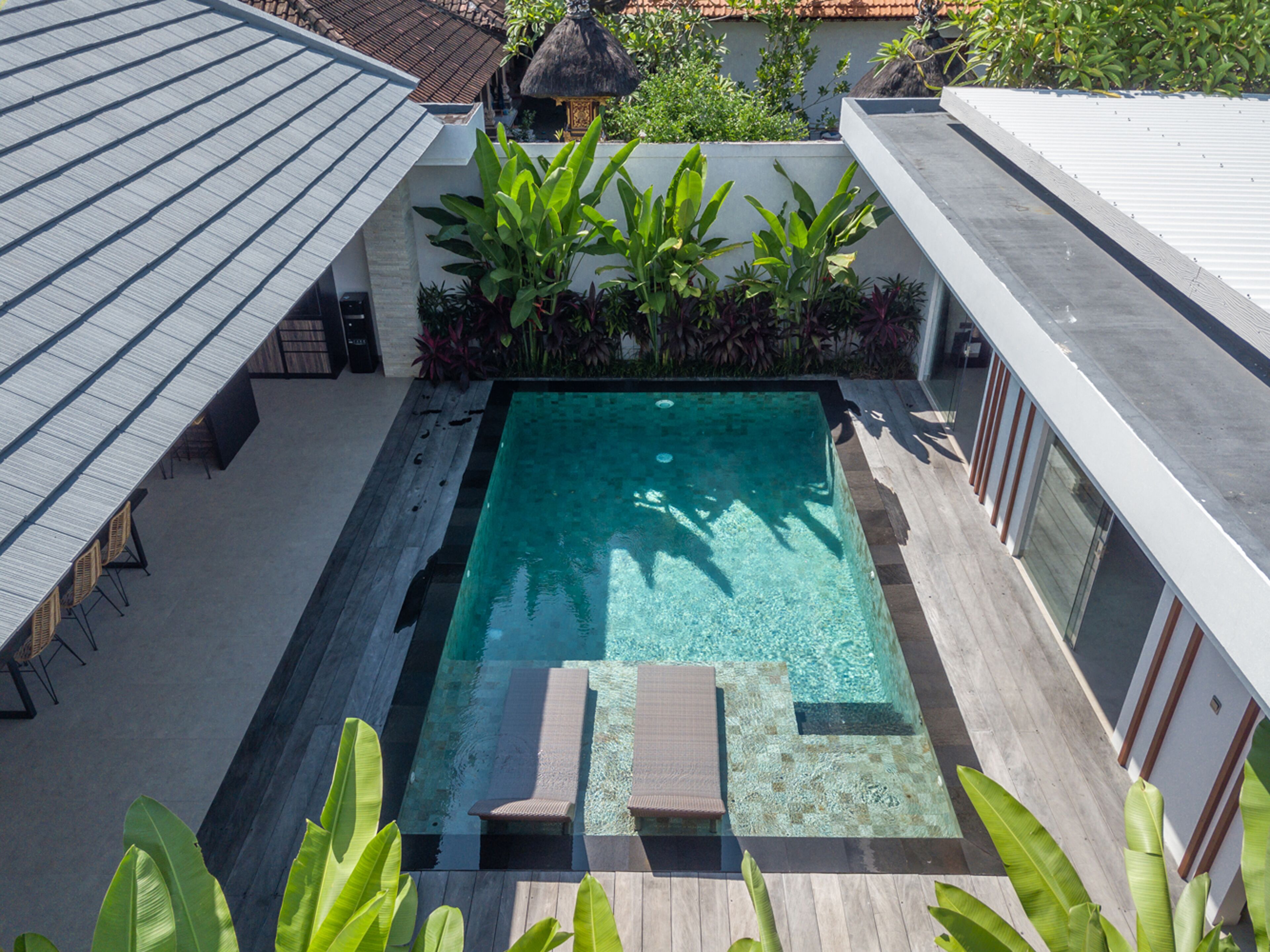 A rectangular swimming pool with a wooden deck and lounge chairs, surrounded by tropical plants and a modern building with a sloped roof.