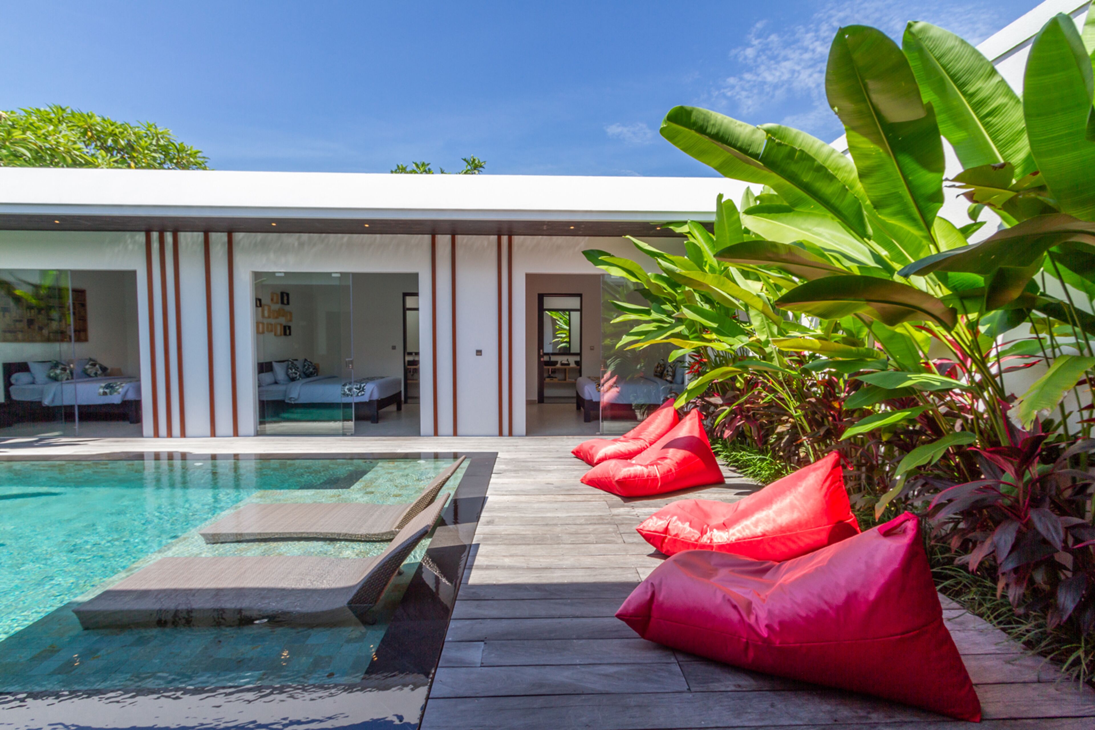 Un área de piscina moderna con una piscina cristalina, terraza de madera y pufs rojos.