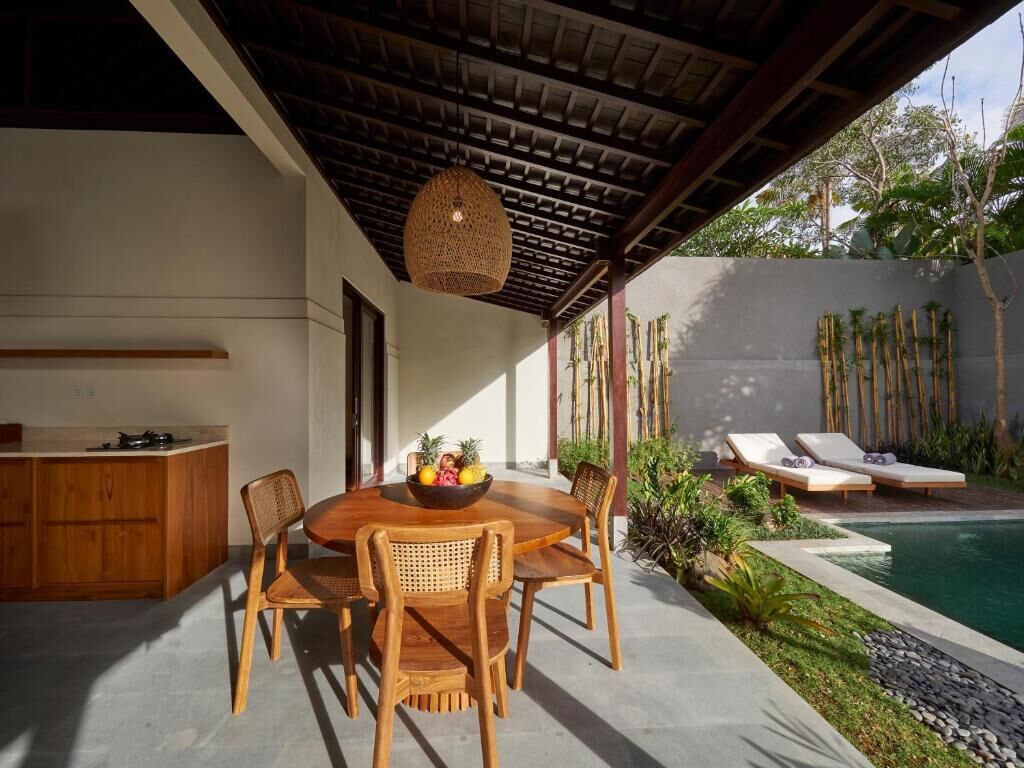 A covered outdoor dining area with a table and chairs, a hanging light fixture, and a poolside lounge area with sun loungers.