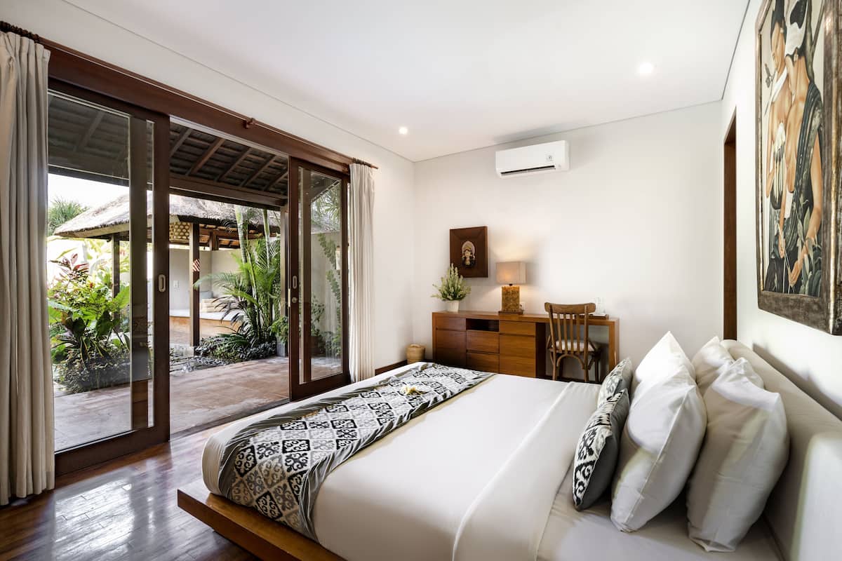 Un dormitorio con una cama, un escritorio y una silla. Hay una puerta corrediza de vidrio que da a un espacio exterior con plantas.