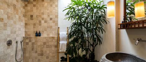 A modern bathroom with a stone sink, a shower with a rain showerhead, a bamboo ladder, and a large potted plant.