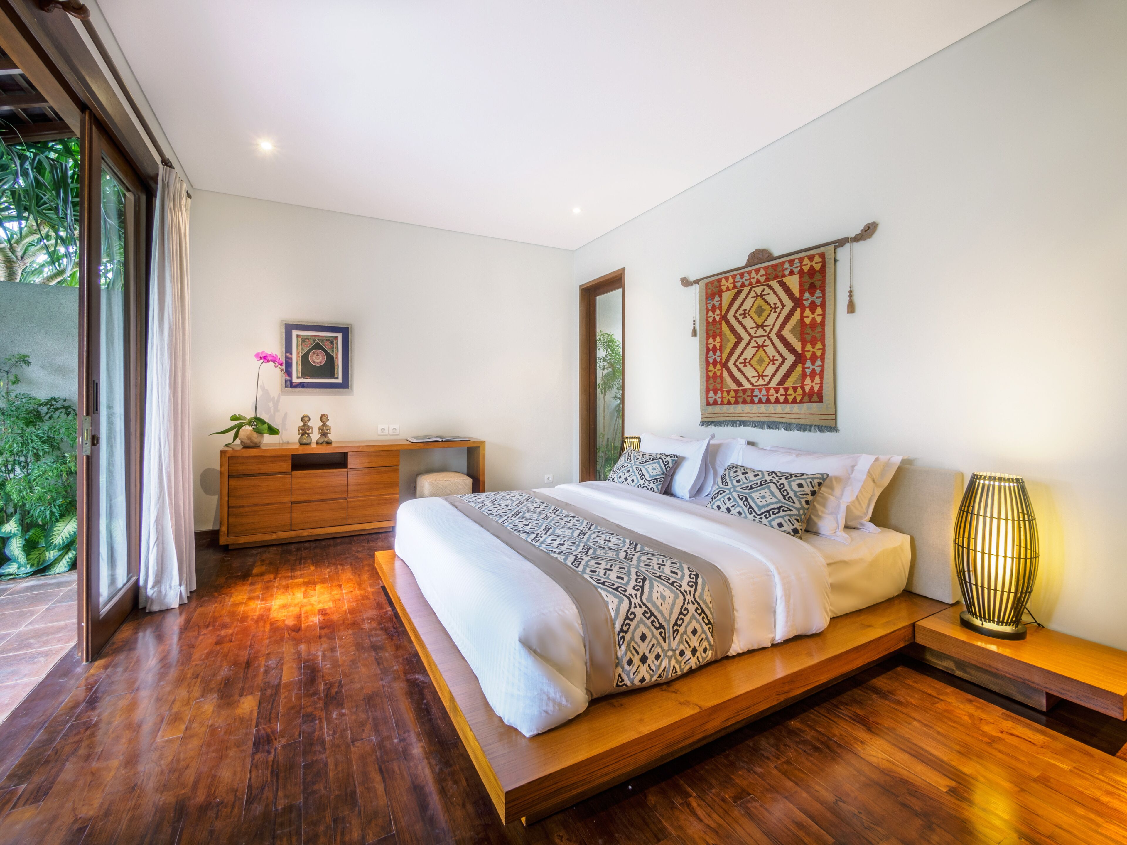 A modern bedroom with a wooden floor, a large bed with patterned bedding, a wooden dresser, and a view of greenery through a sliding door.