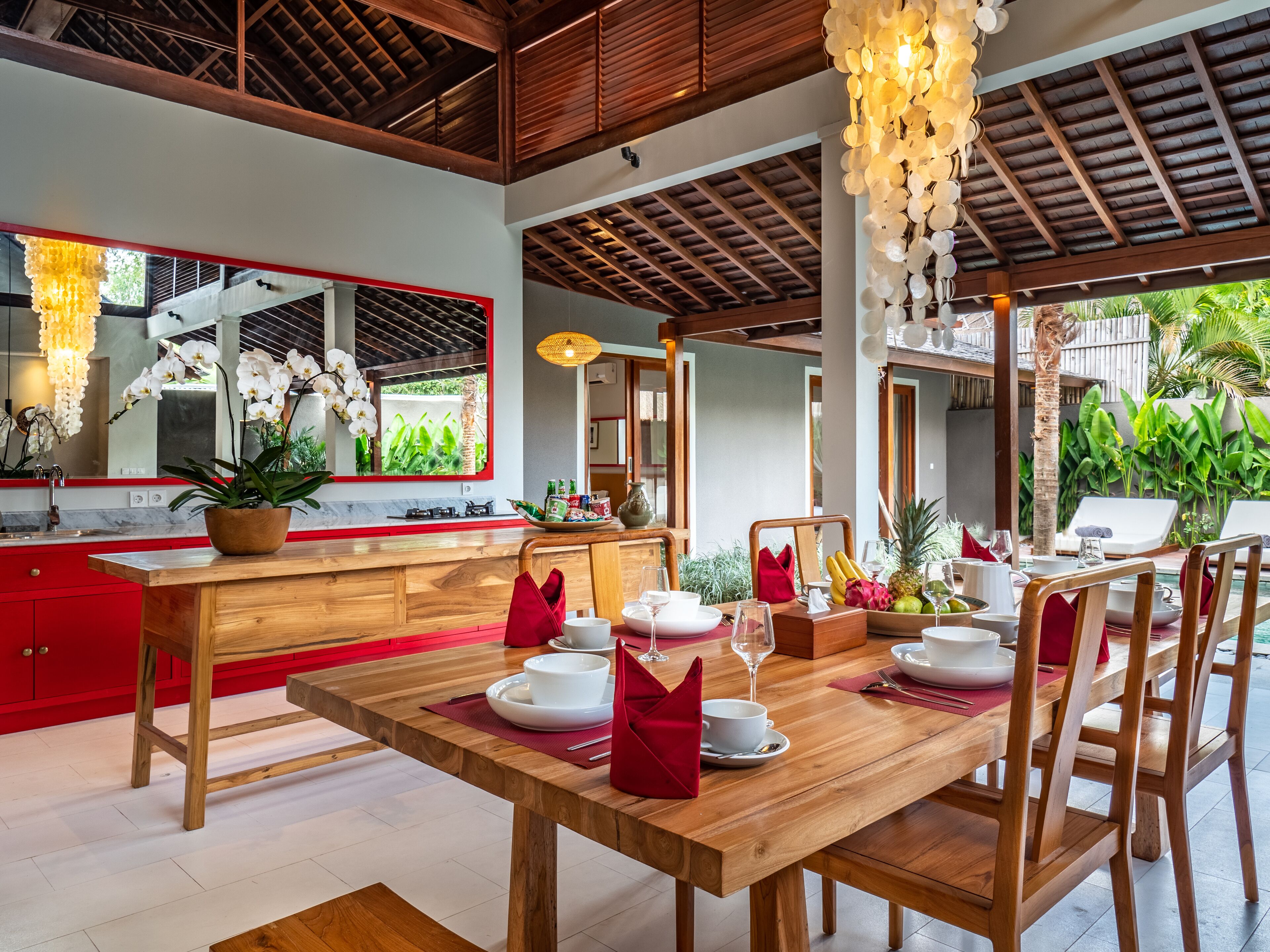 A modern dining area with a wooden table set for four, a kitchen with red cabinets, and a large mirror reflecting the room.