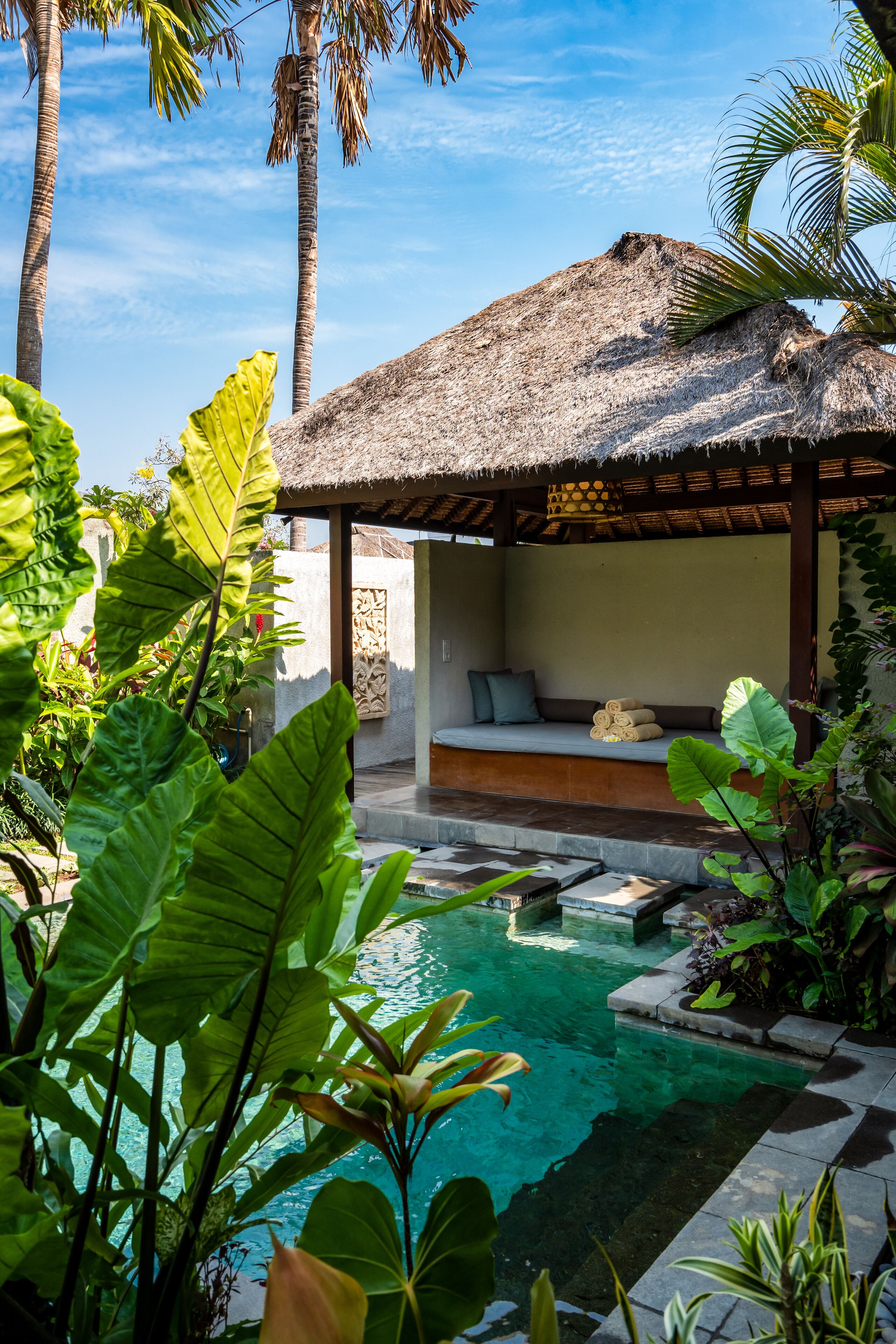 A pool area with a thatched-roof structure, surrounded by lush tropical plants.