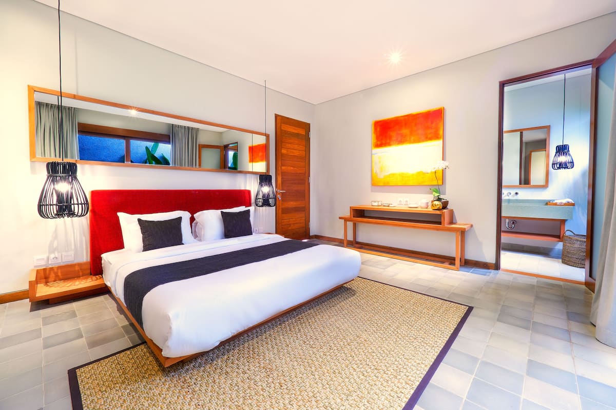 Una habitación de hotel moderna con una cama grande, una mesita de noche de madera, un espejo y un baño a la vista a través de una puerta abierta.