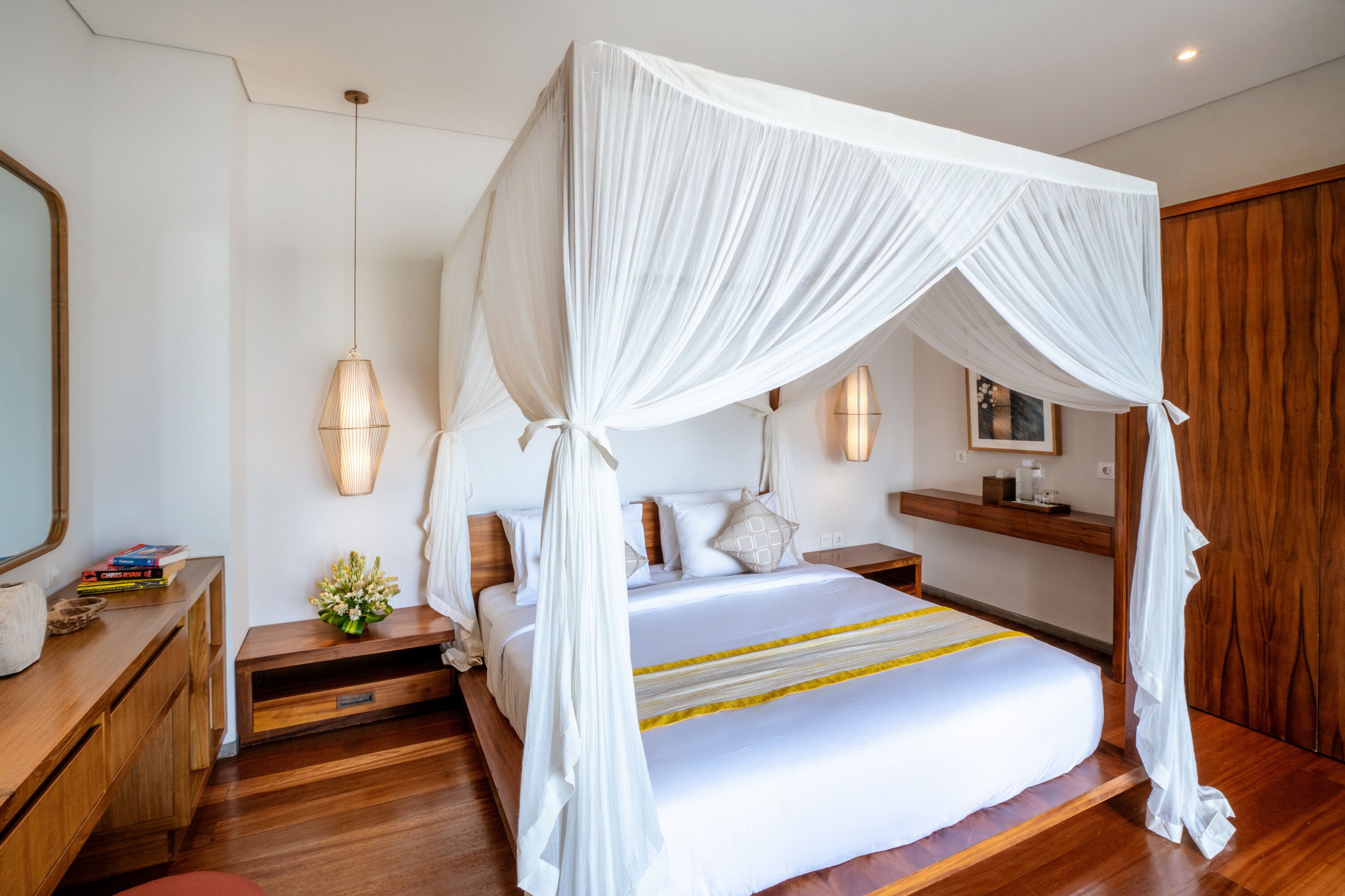 Une chambre à coucher avec un lit à baldaquin, un plancher en bois, des tables de chevet, une commode, un miroir et une étagère fixée au mur.