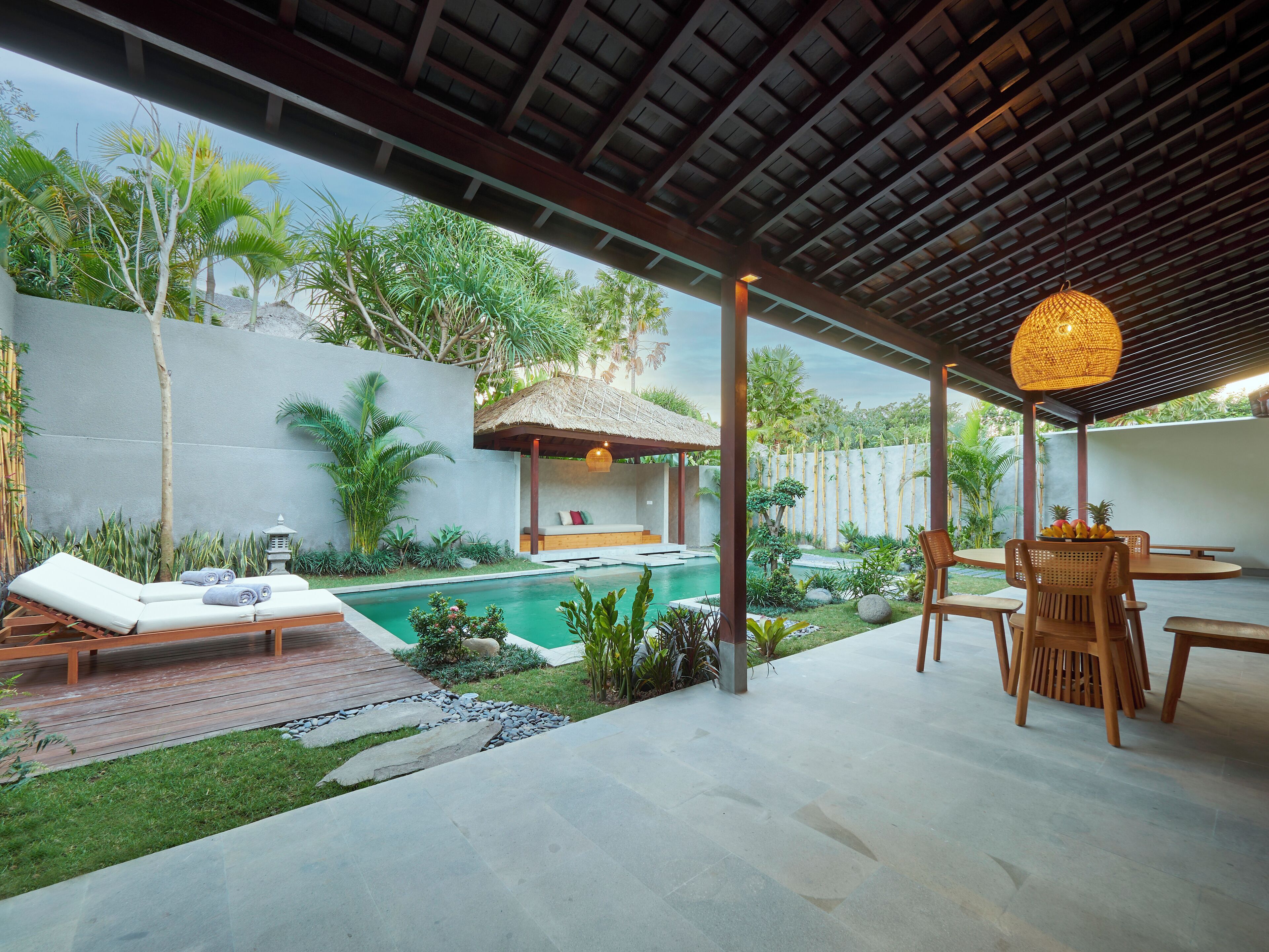 Uma área externa coberta com piscina, espreguiçadeiras, mesa de jantar e uma estrutura com telhado de palha.